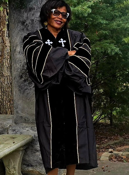 Dr. Dian JacksonDavis standing outdoors in pastoral robes beneath a stone archway, smiling with her arms crossed.